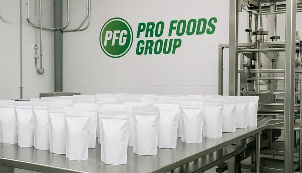 Large order of white stand-up pouches neatly staged on a stainless steel conveyor table inside Pro Foods Group co-packing facility, with company logo displayed on the wall in the background.